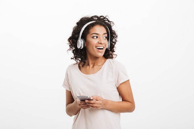 Mulher americana moderna com penteado afro e sorriso grande, olhando de lado enquanto ouve música através de fones de ouvido sem fio e telefone celular, isolado sobre a parede branca