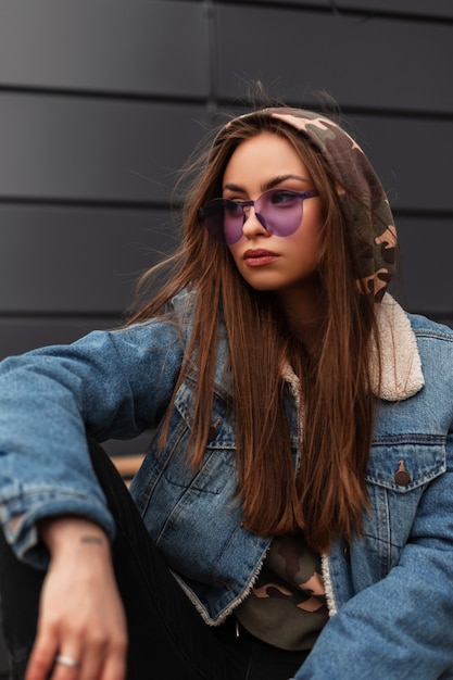 Mulher americana moderna jovem hippie em elegantes óculos roxos com jaqueta jeans azul jovem é relaxante ao ar livre perto de um prédio cinza. Modelo de menina bonita com roupas casuais na rua. Estilo juvenil de primavera.