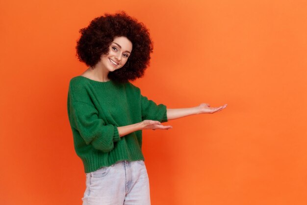 Mulher amigável feliz com penteado afro vestindo suéter verde estilo casual apresentando área de publicidade com espaço de cópia de sorriso dentuço Tiro de estúdio interior isolado em fundo laranja