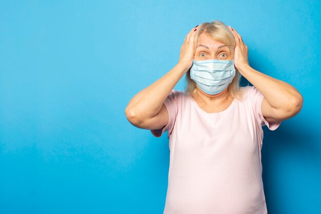 Foto mulher amigável idosa em uma máscara protetora médica segurando as mãos na cabeça com cara de surpresa na parede azul. rosto emocional. vírus, quarentena, pandemia. gesto de ansiedade, preocupação, choque