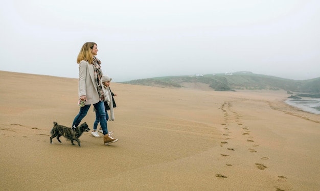 Mulher andando com a filha e o cachorro na praia em um dia nublado de inverno