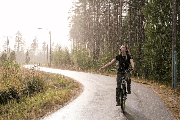 mulher andando de bicicleta ao lado da floresta Ela manteve uma mão no volante sorrindo e aproveitando a chuva quente de verão