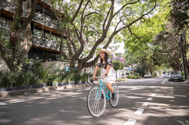 Mulher andando de bicicleta na rua com árvores nas laterais usando vestido e chapéu brancos