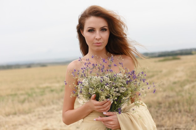 Mulher andando no campo de grama seca dourada Beleza de retrato natural Linda garota em um campo de trigo Jovem mulher em um vestido bege segurando um buquê de flores silvestres