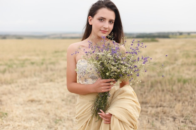 Mulher andando no campo de grama seca dourada beleza de retrato natural linda garota em um campo de trigo jovem mulher em um vestido bege segurando um buquê de flores silvestres