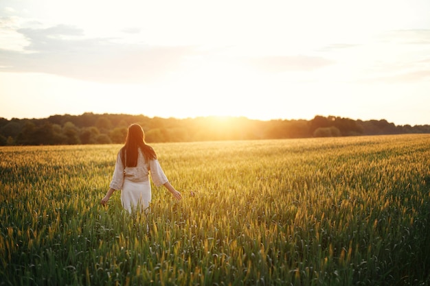 Mulher andando no campo de trigo na luz quente do pôr do sol Elegante jovem mulher em vestido rústico segurando flores silvestres nas mãos e relaxando na paisagem de verão à noite Momento atmosférico tranquilo