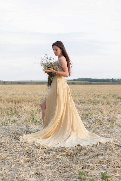 Mulher andando no campo dourado de grama seca. Beleza do retrato natural. Linda garota em um campo de trigo. Jovem mulher com um vestido bege, segurando um buquê de flores silvestres.