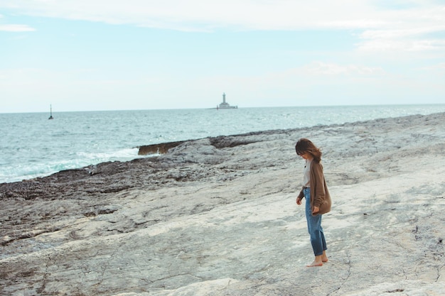 Mulher andando pela praia do mar rochoso no farol de jeans molhado no fundo