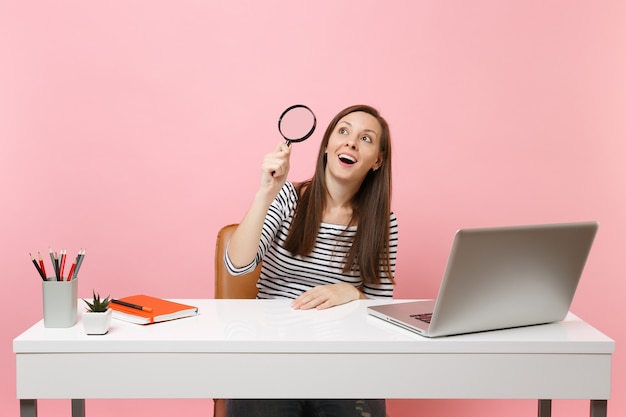 Mulher animada com roupas casuais olhando para cima através da lupa trabalhando em uma mesa branca com um laptop pc contemporâneo