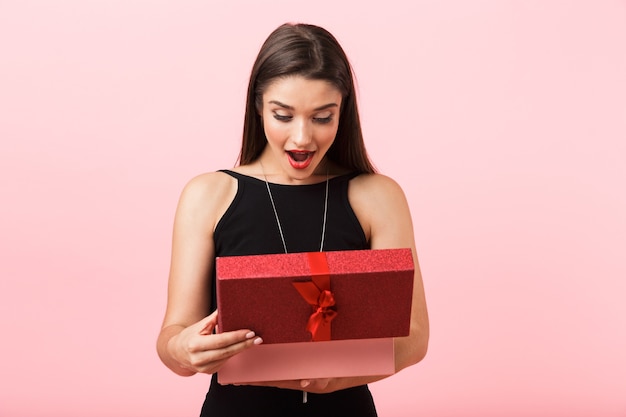 Mulher animada com um vestido preto segurando uma caixa de presente aberta isolada sobre um fundo rosa