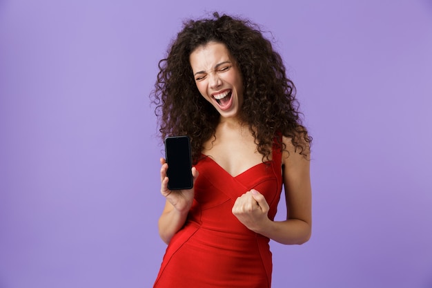 mulher animada com um vestido vermelho segurando um smartphone preto, isolada na parede violeta