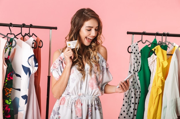 mulher animada em pé perto do guarda-roupa, segurando um smartphone e um cartão de crédito isolado em rosa