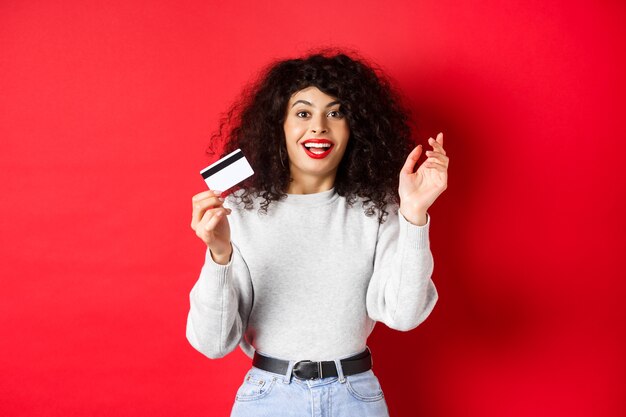 Mulher animada, falando sobre uma oferta especial, mostrando o cartão de plástico e parecendo feliz, indo às compras, em pé sobre fundo vermelho.