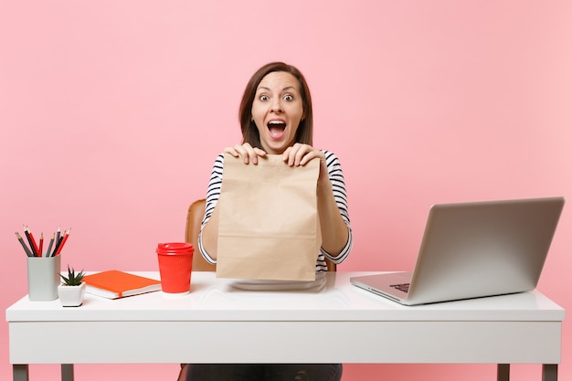 Mulher animada segurando na mão um saco de papel artesanal em branco transparente marrom trabalhando no escritório com o laptop