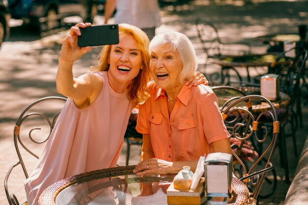 Mulher animada segurando um smartphone e tirando uma selfie com a mãe idosa