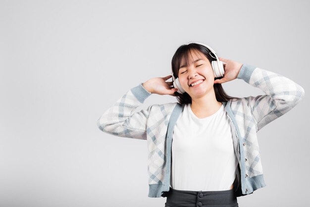 Mulher animada sorrindo ouvindo rádio de música em fones de ouvido bluetooth e segurando um tiro de estúdio de telefone fundo branco isolado, feliz jovem asiática ouve telefone móvel de tecnologia moderna