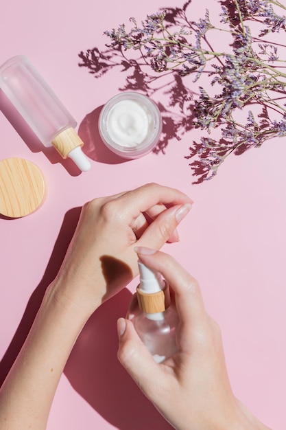 Mulher aplicando creme na mão com fundo rosa com produtos cosméticos e flores Conceito de tratamento de cuidados com a pele