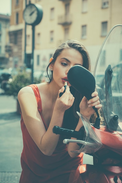 Mulher aplicando maquiagem na rua. Modelo de menina moda sexy com lábios vermelhos passar batom olhando em uma motocicleta espelho.