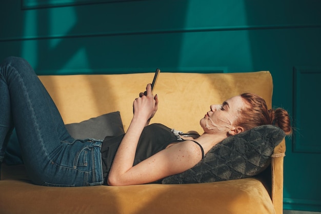 Mulher aplicando máscara e usando telefone celular deitado no sofá em casa cuidados com a pele rosto feliz tre facial