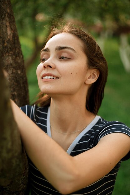 Mulher apoiando-se no galho de árvore e aproveitando o verão em pé no jardim verde