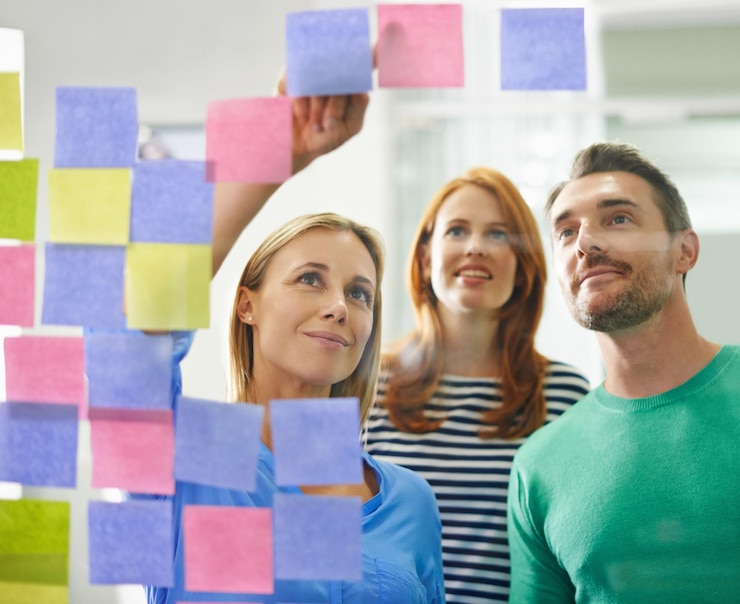 Foto mulher aponta e anota em vidro com trabalho em equipe para planejar revisão e feliz por ideias em escritório moderno pessoas de negócios moodboard e sorrir com homem grupo e pensar em startup de mídia criativa