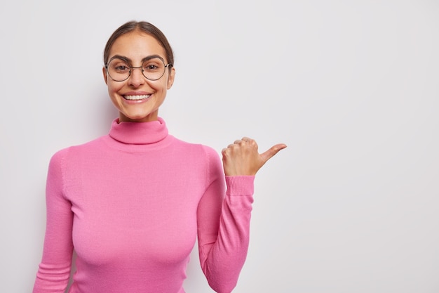 Mulher aponta o polegar para longe anuncia que seu produto promove venda ou desconto vestido em shows casuais de gola alta rosa