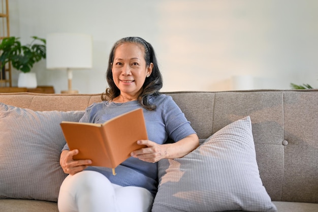 Mulher aposentada de idade asiática feliz lendo um livro no sofá em sua sala de estar aconchegante mínima