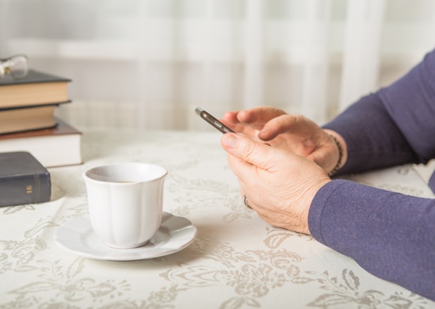 Mulher aposentada está usando telefone celular, na acolhedora cozinha em casa. Avó tem café na cozinha