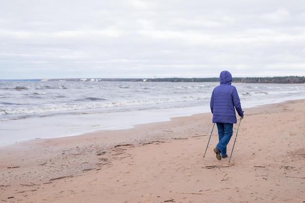 Mulher aposentada sênior caminhando à beira-mar na areia Aptidão ativa e estilo de vida saudável Treinamento feminino na praia Caminhada nórdica com bastões Temporada de outono à beira-mar Casal esportivo