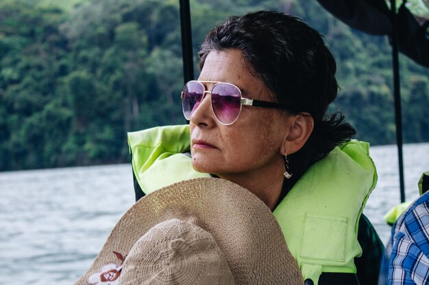 mulher aposentada sênior com óculos e chapéu com colete salva-vidas navegando em um rio em um barco de férias