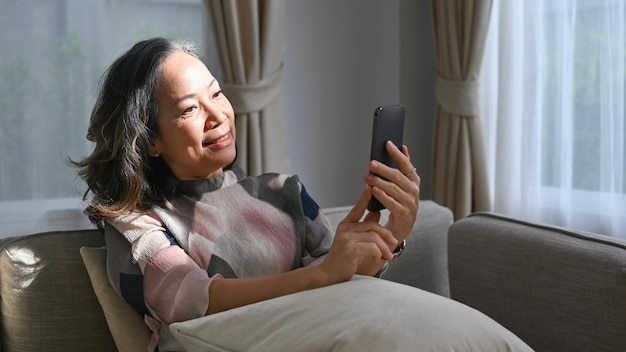 Mulher aposentada sorridente usando smartphone no sofá em casa Conceito de tecnologia de aposentadoria
