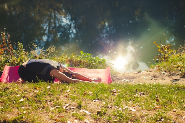 Mulher apta saudável, estendendo-se no tapete de ioga rosa à beira do rio no parque da cidade de verão. Moça que se estende na grama verde à beira do rio com o sol refletido na água