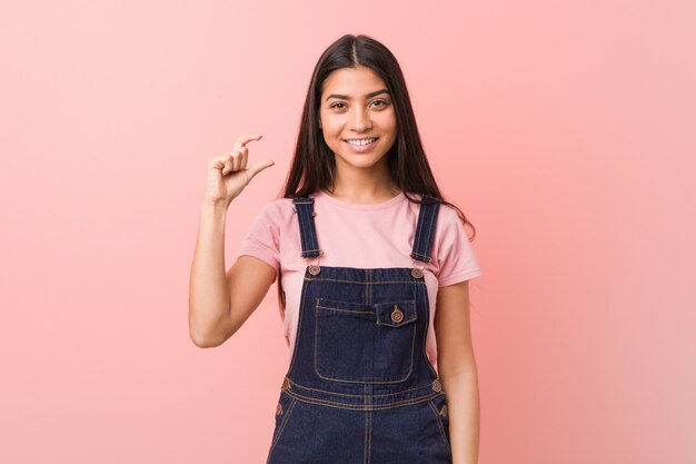 Mulher árabe bonita nova que veste um dungaree das calças de brim que guarda algo pequeno com os dedos indicadores, sorrindo e confiantes.