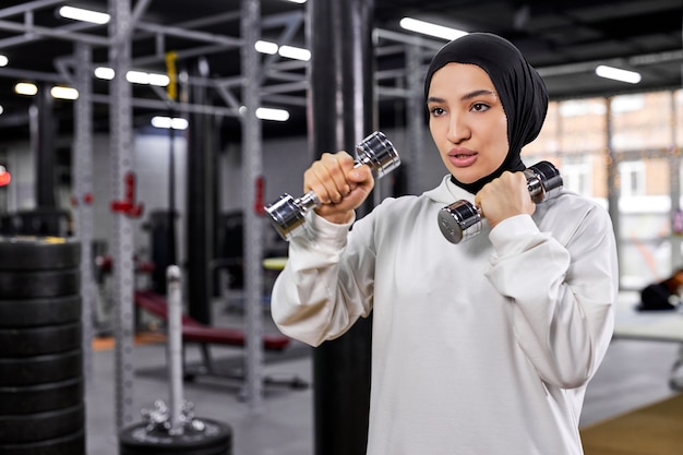 Mulher árabe em hijab branco, exercitando-se com halteres no ginásio, desfrutando de um estilo de vida saudável e ativo, concentrado no treino, copie o espaço. esporte, fitness, conceito de treinamento