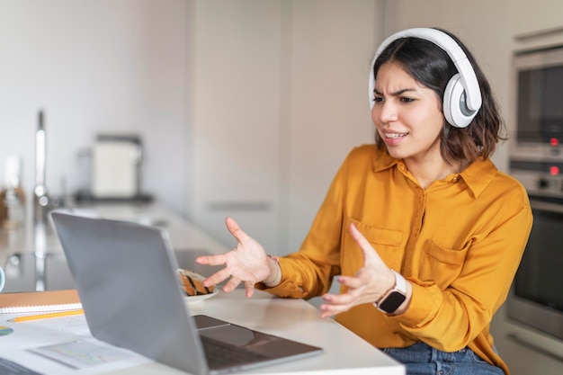 Mulher árabe estressada em fones de ouvido fazendo videochamada no laptop na cozinha