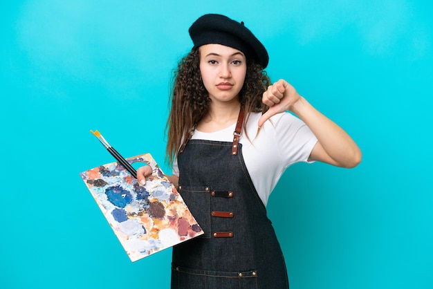 Mulher árabe jovem artista segurando uma paleta isolada em fundo azul, mostrando o polegar para baixo com expressão negativa