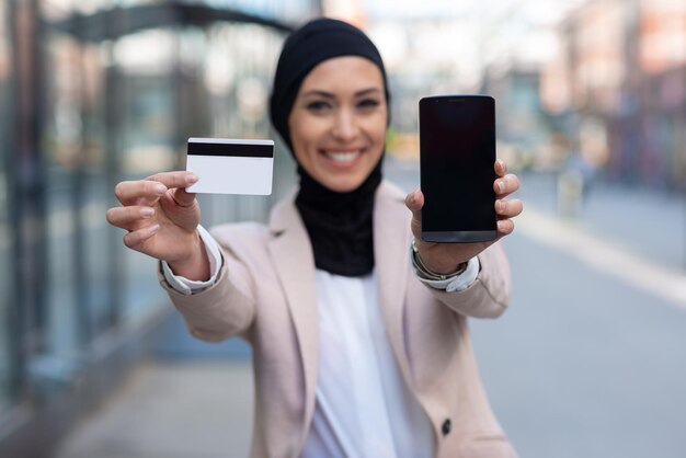 Mulher árabe segurando cartão de crédito e telefone ao ar livreFeche o retrato da mulher muçulmana segurando o telefone