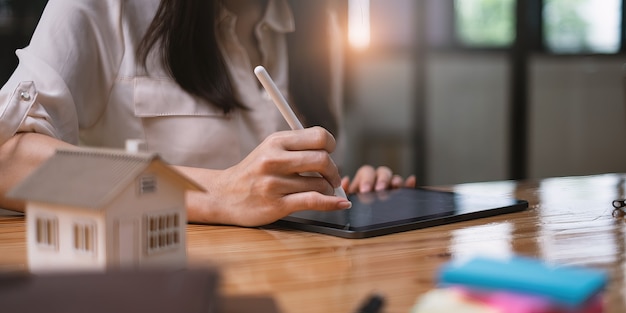 Mulher arquiteta usando um aplicativo de tablet digital para desenhar à mão uma casa de projeto com um modelo em uma mesa de madeira. Ideia de propriedade no mercado imobiliário.