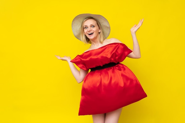 Mulher arrasadora e animada em um travesseiro vermelho e chapéu de verão estende os braços para o lado em estúdio amarelo