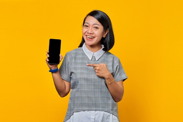 Mulher asiática alegre mostrando a tela do smartphone em branco e apontando para ela enquanto olha para a câmera sobre fundo amarelo