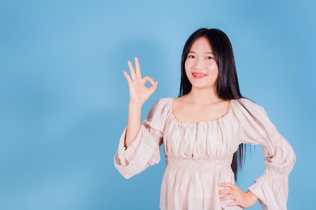 Mulher asiática alegre sorridente usa camiseta branca mostrando oksign em fundo azul