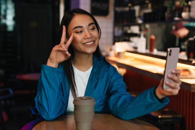 Mulher asiática alegre tirando uma selfie no celular e gesticulando o sinal da paz enquanto está sentada no café