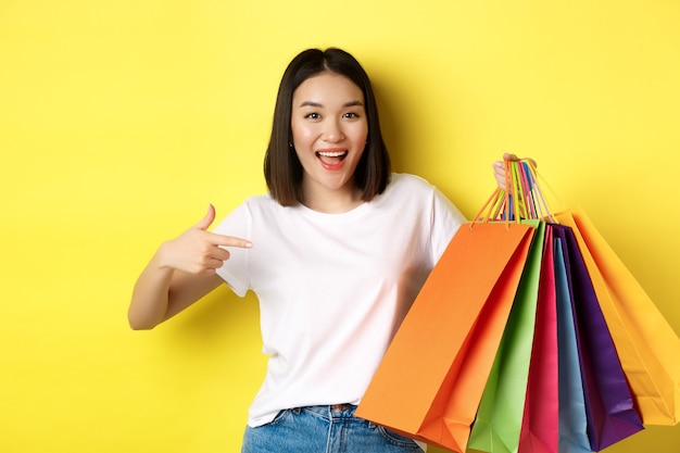 Mulher asiática alegre vai às compras, apontando o dedo para sacos de papel e sorrindo