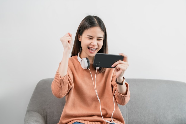 Mulher asiática animada jogando jogos no smartphone na sala de estar