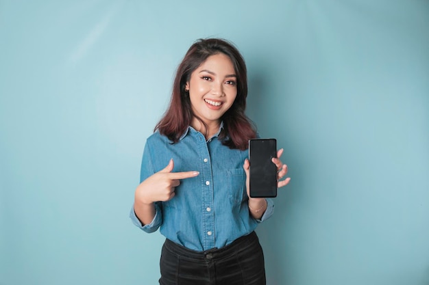 Mulher asiática animada vestindo camisa azul apontando para o espaço de cópia em seu smartphone isolado por fundo azul
