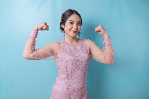 Mulher asiática animada vestindo roupas modernas de kebaya mostrando um gesto forte levantando seus braços e músculos sorrindo orgulhosamente