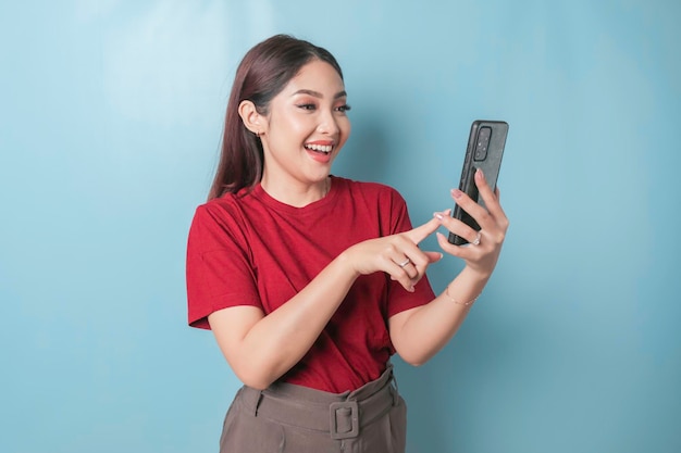 Mulher asiática animada vestindo uma camiseta vermelha apontando para seu smartphone isolado por um fundo azul
