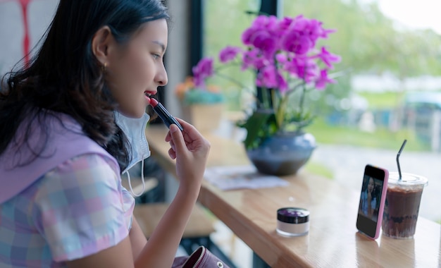 Mulher asiática aplicar batom vermelho nos lábios em um café usando o smartphone como espelho, garota tomar uma bebida e se maquiar na mesa de madeira no café