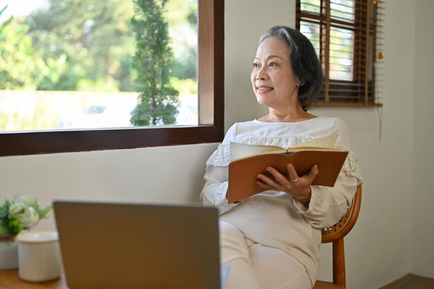 Mulher asiática aposentada relaxada dos anos 60 olhando pela janela sonhando acordada enquanto lê um livro