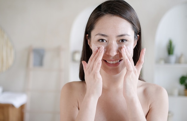 Foto mulher asiática atraente aplicar creme para a pele no rosto olhar no espelho do banheiro em casa jovem mestiça feliz colocar creme facial hidratante para levantamento de pele e conceito de tratamento corporal saudável